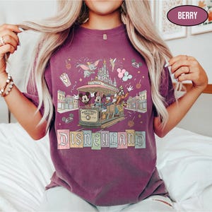 May include: Berry-colored t-shirt featuring a Disneyland Main Street Trolley design with Mickey, Minnie, Donald, and Goofy. The shirt includes the Disneyland logo and other park icons like Dumbo and fireworks.
