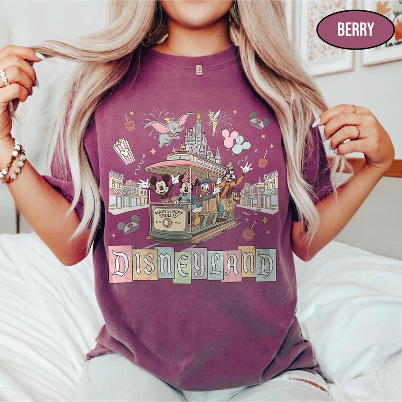 May include: Berry-colored t-shirt featuring a Disneyland Main Street Trolley design with Mickey, Minnie, Donald, and Goofy. The shirt includes the Disneyland logo and other park icons like Dumbo and fireworks.