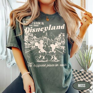 May include: Moss green t-shirt with a Disneyland graphic. The design features Mickey and Minnie Mouse, the text "California Est. 1955 Disneyland", and "The happiest place on earth". The shirt has a vintage, distressed look.