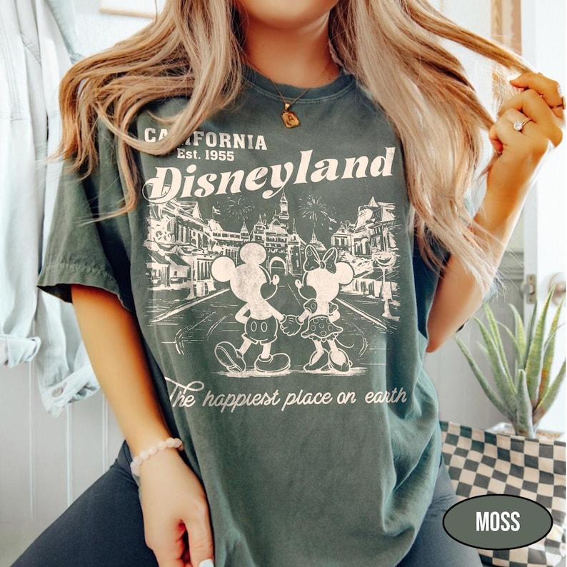 May include: Moss green t-shirt with a Disneyland graphic. The design features Mickey and Minnie Mouse, the text "California Est. 1955 Disneyland", and "The happiest place on earth". The shirt has a vintage, distressed look.