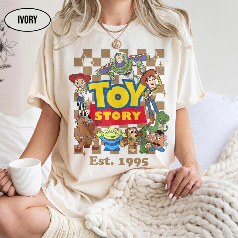 May include: Ivory-colored t-shirt featuring a graphic of Toy Story characters, including Woody, Buzz Lightyear, and others, with the words "TOY STORY" and "Est. 1995". The design is set against a brown and white checkered background.