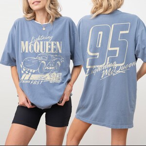 Puede incluir: Camiseta azul claro con gr&aacute;ficos de Lightning McQueen. La parte delantera muestra el texto "Lightning McQueen" encima de una ilustraci&oacute;n de coche y "THINK FAST 95". La parte trasera tiene un gran "95" y el texto "Lightning McQueen". La camiseta se usa con pantalones cortos negros.