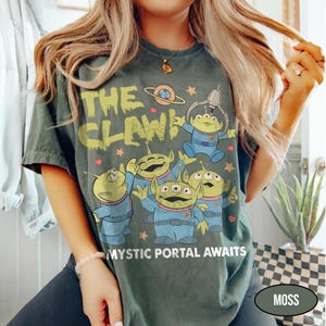 May include: Moss green t-shirt featuring the text "THE CLAW!" above a graphic of green aliens in blue suits. The phrase "MYSTIC PORTAL AWAITS" is printed below the aliens. The shirt has a vintage, worn look.