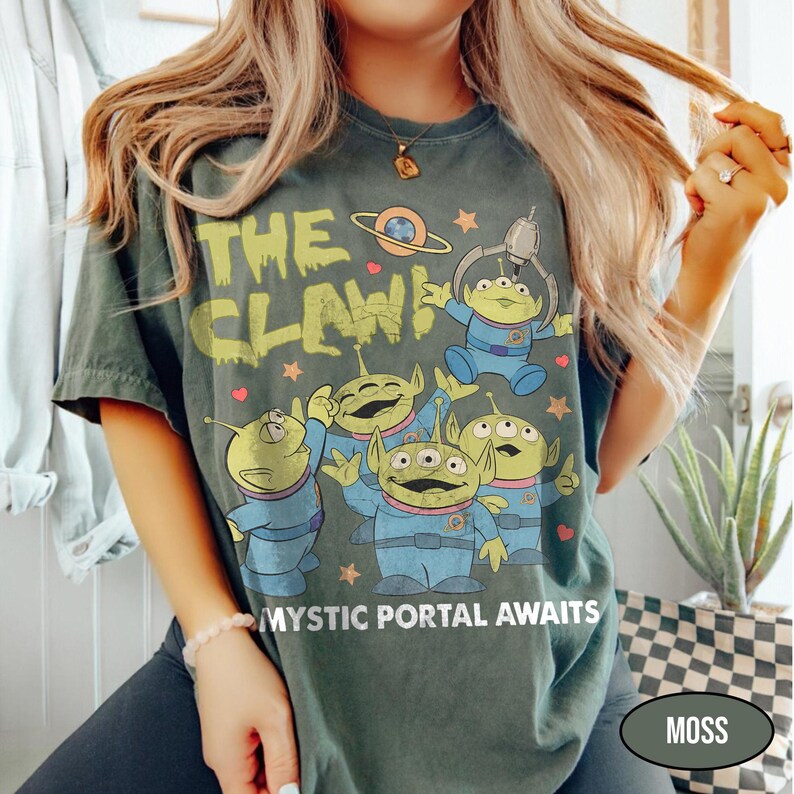 May include: Moss green t-shirt featuring the text "THE CLAW!" above a graphic of green aliens in blue suits. The phrase "MYSTIC PORTAL AWAITS" is printed below the aliens. The shirt has a vintage, worn look.
