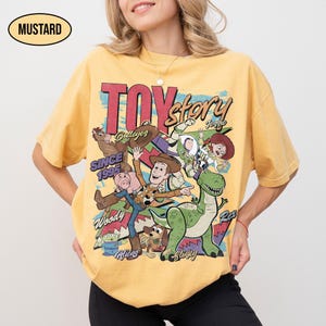 Puede incluir: Camiseta de color mostaza con un gr&aacute;fico de personajes de la pel&iacute;cula de animaci&oacute;n "Toy Story". El dise&ntilde;o incluye a Woody, Buzz Lightyear, Jessie, Rex y otros personajes, con el texto "TOY STORY" y "SINCE 1995".