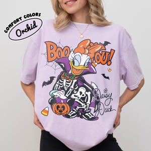 May include: A light purple t-shirt featuring a graphic of Daisy Duck dressed in a skeleton costume with a cape, holding a pumpkin. The text reads Boo You and Daisy Duck, surrounded by spiderwebs, bats, and candy corn. A label reads Comfort Colors Orchid.