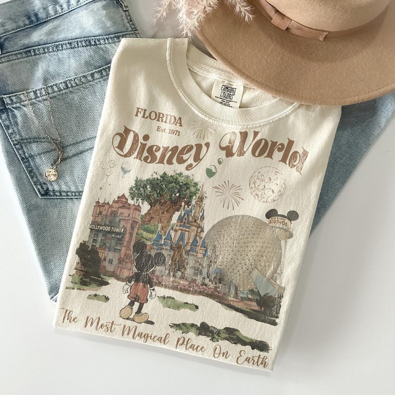 Puede incluir: Camiseta crema de Disney World con un gr&aacute;fico de estilo vintage que presenta a Mickey Mouse, el castillo de Cenicienta y la Torre Hollywood. La camiseta incluye el texto "Florida Est. 1971" y "The Most Magical Place On Earth."