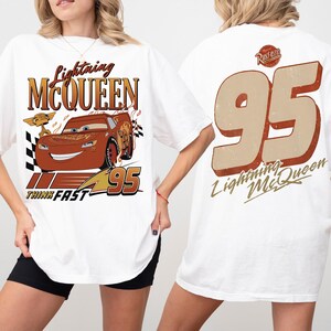 May include: White t-shirt with a Lightning McQueen design. The front shows the character, the text "Lightning McQueen", and "95". The back has a large "95" and "Lightning McQueen" text. A fun, casual tee.