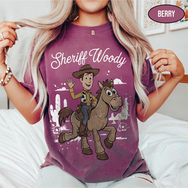 May include: A berry-colored t-shirt featuring a graphic of Sheriff Woody riding a brown horse. The text "Sheriff Woody" is printed in white script above the characters. The design includes a desert scene with a cactus and a skull in white.