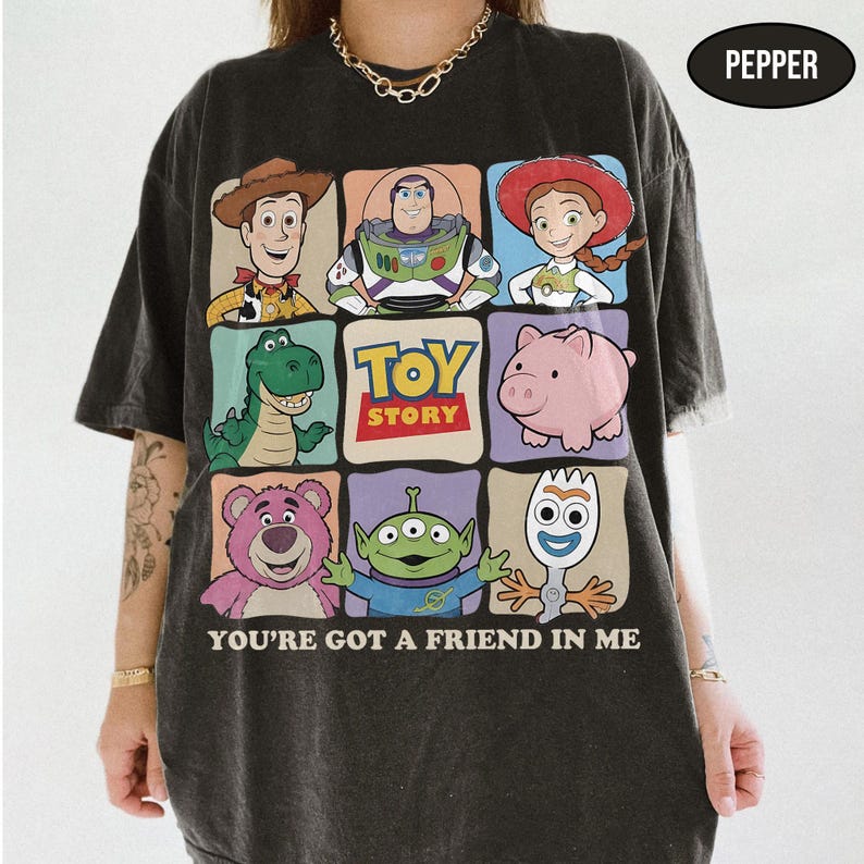 May include: Dark gray t-shirt featuring a colorful graphic of Toy Story characters. The design includes Woody, Buzz Lightyear, Jessie, Rex, Hamm, Lotso, an alien, and Forky. The text "TOY STORY" and "YOU'RE GOT A FRIEND IN ME" are also displayed.