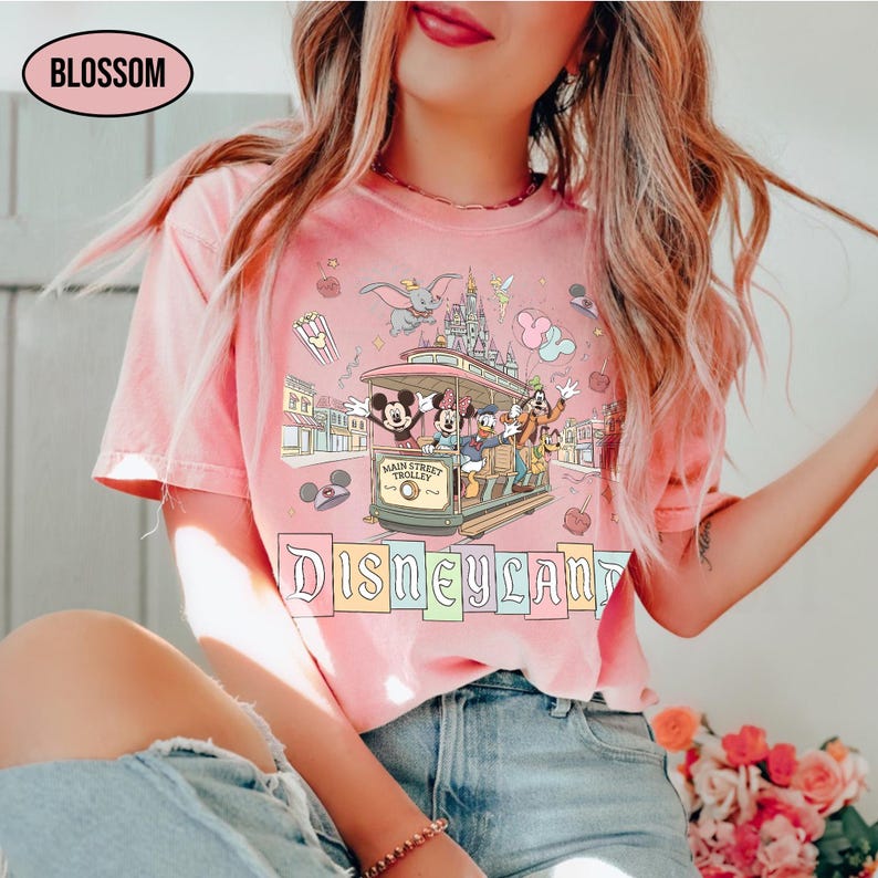 May include: Pink t-shirt featuring a Disneyland design with Mickey Mouse, Minnie Mouse, Donald Duck, Goofy, and other characters riding a Main Street Trolley. The word "Disneyland" is displayed in colorful block letters. The shirt also has the word "Blossom" in a black oval.