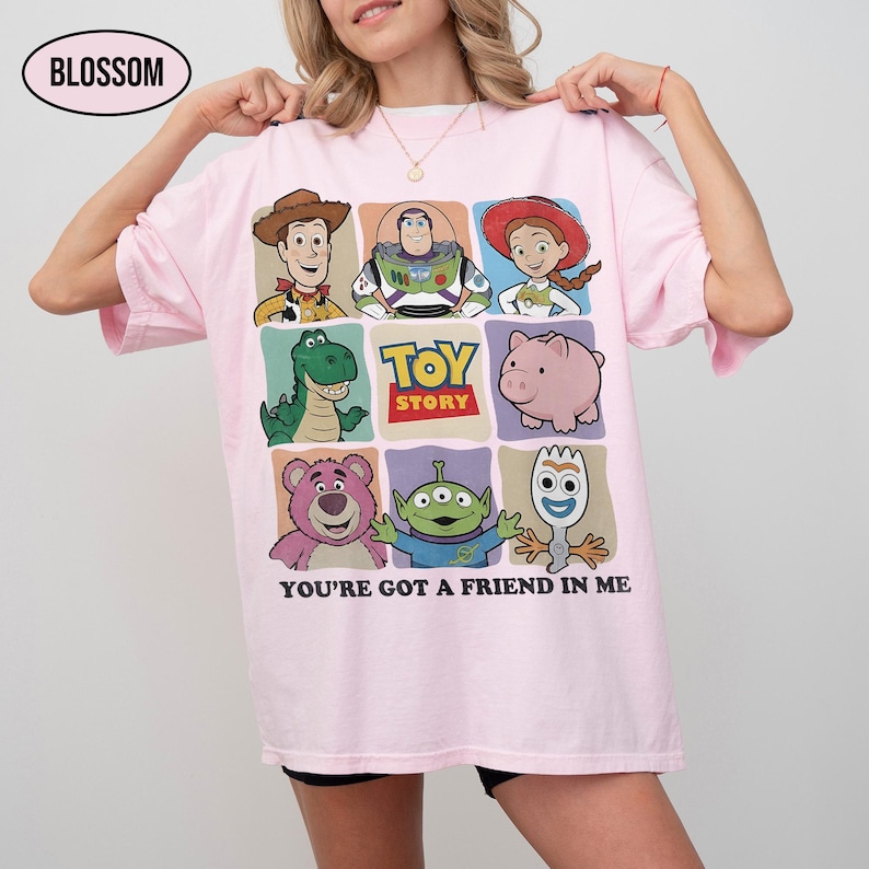May include: Light pink t-shirt featuring a colorful graphic of Toy Story characters. The design includes Woody, Buzz Lightyear, Jessie, Rex, Hamm, Lotso, the Aliens, and Forky. The text "TOY STORY" and "YOU'RE GOT A FRIEND IN ME" are also displayed.