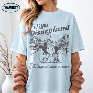 May include: Light blue t-shirt featuring a black and white graphic of Mickey and Minnie Mouse holding hands in front of a Disneyland scene. The shirt includes the text "California Disneyland Est. 1955" and "The happiest place on earth."