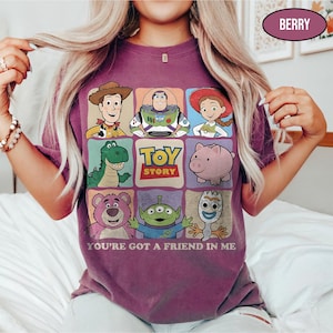 May include: Berry-colored t-shirt featuring a grid of Toy Story characters including Woody, Buzz Lightyear, and Jessie. The shirt has the text "TOY STORY" and "YOU'RE GOT A FRIEND IN ME" printed on it. The shirt is a short-sleeved crew neck.