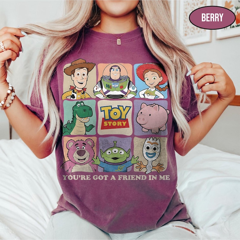 May include: Berry-colored t-shirt featuring a grid of Toy Story characters including Woody, Buzz Lightyear, and Jessie. The shirt has the text "TOY STORY" and "YOU'RE GOT A FRIEND IN ME" printed on it. The shirt is a short-sleeved crew neck.