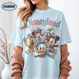 May include: Light blue t-shirt featuring the word "Disneyland" in a colorful, retro font. The shirt showcases a group of Disney characters, including Mickey Mouse, Minnie Mouse, Donald Duck, Goofy, and Pluto, in a selfie-style design.