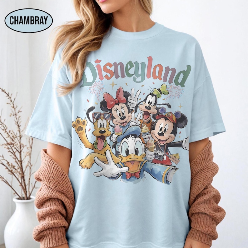 May include: Light blue t-shirt featuring the word "Disneyland" in a colorful, retro font. The shirt showcases a group of Disney characters, including Mickey Mouse, Minnie Mouse, Donald Duck, Goofy, and Pluto, in a selfie-style design.