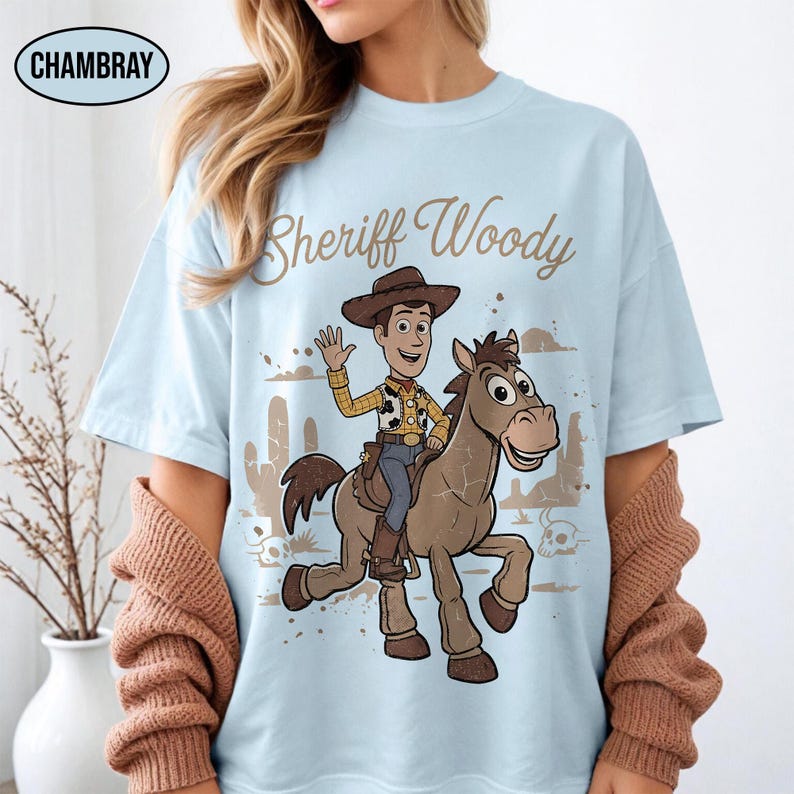 May include: A light blue t-shirt featuring a graphic print of Sheriff Woody riding a brown horse. The text "Sheriff Woody" is printed in brown script above the character. The design includes desert elements like cacti and skulls in a muted brown tone.