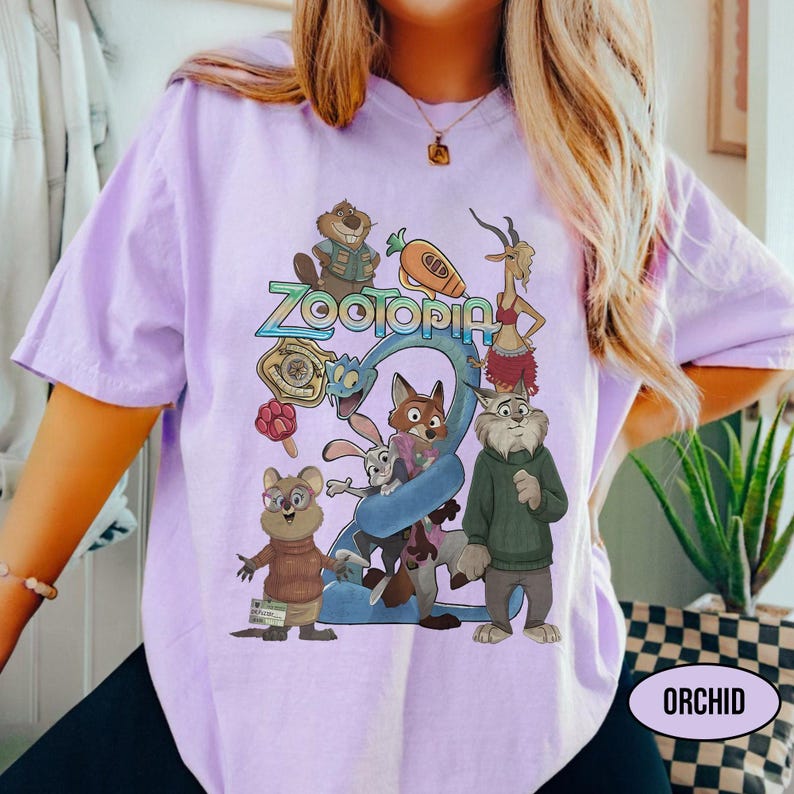 May include: A light purple t-shirt featuring a colorful graphic of characters from the animated movie "Zootopia". The design includes the word "Zootopia" in a stylized font, along with various animal characters. The shirt has short sleeves and a relaxed fit.