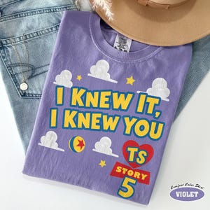 Puede incluir: Una camiseta violeta con el texto amarillo I KNEW IT, I KNEW YOU, gr&aacute;ficos de nubes blancas, una pelota amarilla y roja, un coraz&oacute;n rojo con las letras TS y el texto rojo STORY 5. La camiseta tiene una etiqueta Comfort Colors de talla XL.