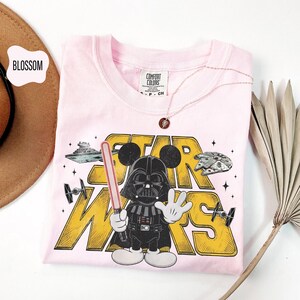 Puede incluir: Una camiseta rosa claro con un gr&aacute;fico de Darth Vader con orejas de Mickey Mouse, sosteniendo un sable de luz, y las palabras "STAR WARS" en amarillo. Tambi&eacute;n se ve un collar de oro rosa y un sombrero marr&oacute;n.
