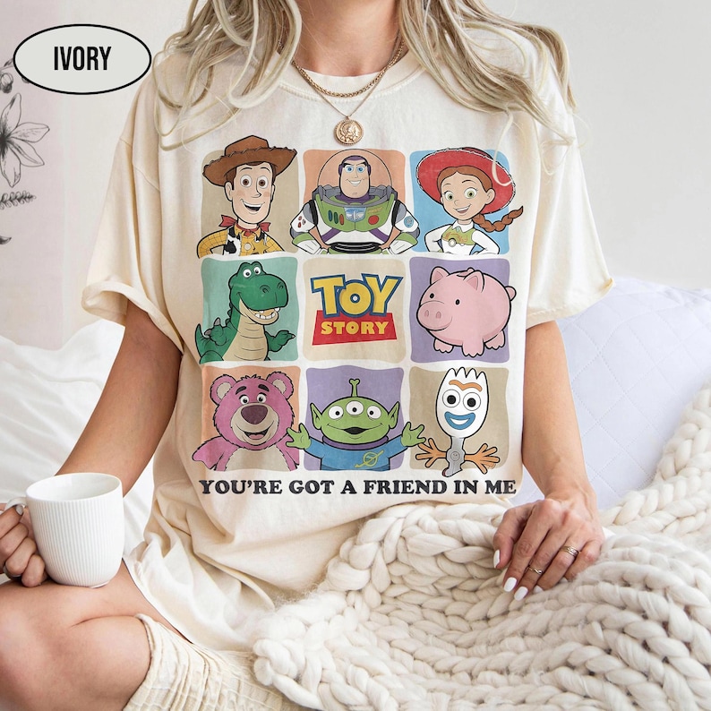 May include: Ivory-colored t-shirt featuring a colorful graphic of Toy Story characters. The design includes Woody, Buzz Lightyear, Jessie, Rex, Hamm, Lotso, the Aliens, and Forky. The text "TOY STORY" and "YOU'RE GOT A FRIEND IN ME" are also displayed.