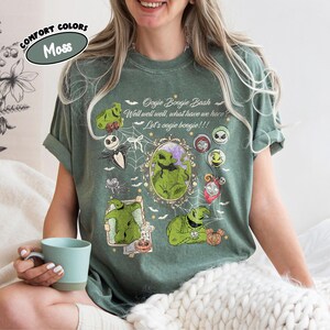 May include: A moss green t-shirt featuring the text "Oogie Boogie Bash" and various cartoon character graphics. The shirt has rolled-up sleeves and a relaxed fit. The shirt also has the text "Well well well, what have we here? Let's oogie boogie!!!"