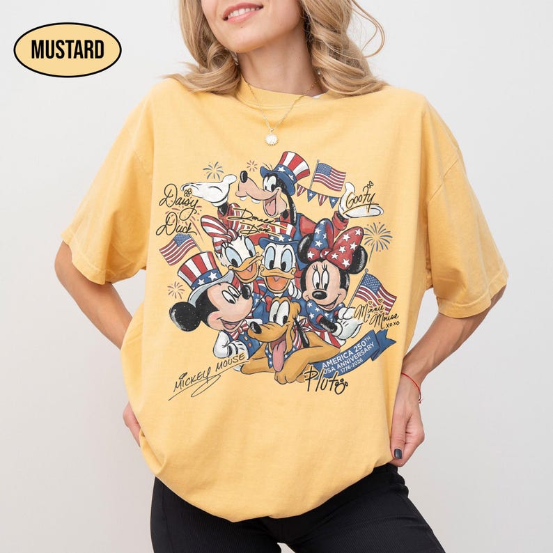 Może przedstawiać: Musztardowa koszulka z grafiką Myszki Miki, Myszki Minnie, Kaczora Donalda, Kaczki Daisy, Goofy'ego i Plutona w patriotycznych strojach, trzymających amerykańskie flagi. Koszulka ma napis "America 250th Anniversary 1776-2026".