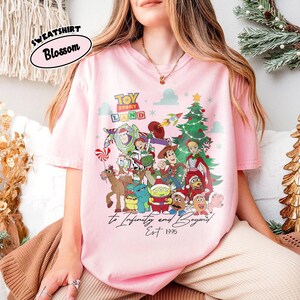 May include: A light pink t-shirt featuring a colorful graphic of Toy Story characters, a Christmas tree, and the text "TOY STORY LAND" and "To Infinity and Beyond Est. 1995". The shirt has short sleeves and a crew neck.