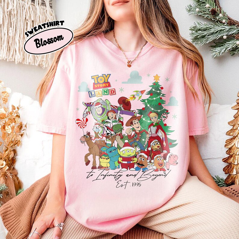 May include: A light pink t-shirt featuring a colorful graphic of Toy Story characters, a Christmas tree, and the text "TOY STORY LAND" and "To Infinity and Beyond Est. 1995". The shirt has short sleeves and a crew neck.