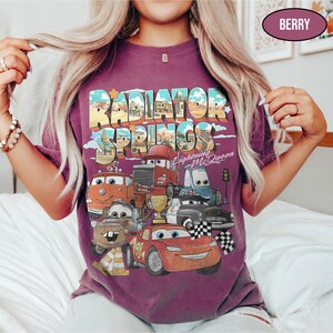 May include: A berry-colored t-shirt featuring a graphic print of Radiator Springs with Lightning McQueen and other characters from the movie. The design includes the text Radiator Springs and Lightning McQueen, along with a gold trophy and checkered flags.