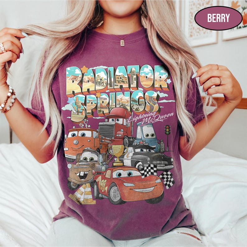 May include: A berry-colored t-shirt featuring a graphic print of Radiator Springs with Lightning McQueen and other characters from the movie. The design includes the text Radiator Springs and Lightning McQueen, along with a gold trophy and checkered flags.