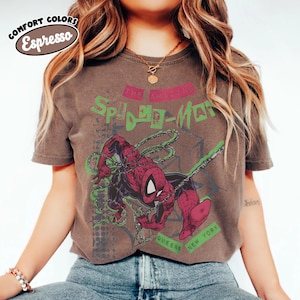 May include: Espresso-colored t-shirt featuring a graphic of Spider-Man in a dynamic pose with web lines. The design includes the text "THE AMAZING SPIDER-MAN" in green and pink, with "QUEENS NEW YORK" below. The shirt has a vintage, distressed look.