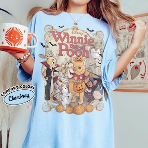 May include: A light blue t-shirt featuring a Disney Winnie the Pooh Halloween graphic with characters dressed in costumes, pumpkins, bats, and spiderwebs. The text reads Disney Winnie the Pooh, Candy, and Mr. Sanders 100 Acre Wood. Comfort Colors Chambray.