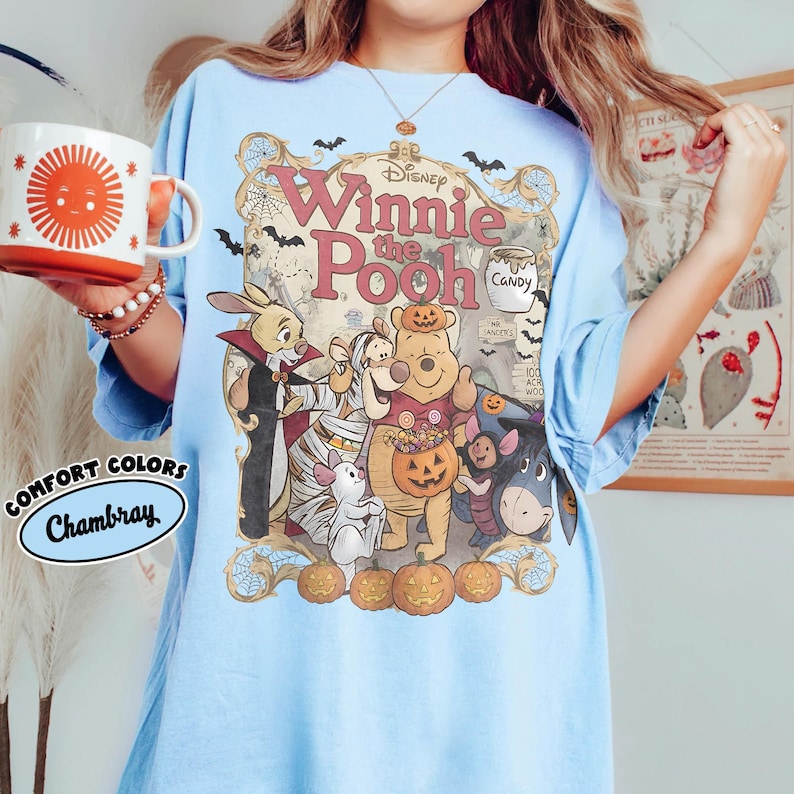 May include: A light blue t-shirt featuring a Disney Winnie the Pooh Halloween graphic with characters dressed in costumes, pumpkins, bats, and spiderwebs. The text reads Disney Winnie the Pooh, Candy, and Mr. Sanders 100 Acre Wood. Comfort Colors Chambray.