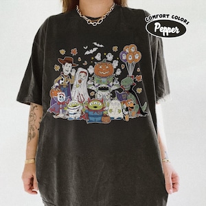 May include: Charcoal grey t-shirt with a Halloween cartoon graphic. The design features characters like Woody, a bride, a pumpkin, and aliens, along with bats, ghosts, and balloons. The text "Comfort Colors Pepper" is printed on the upper right.