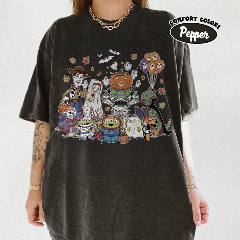 May include: Charcoal grey t-shirt with a Halloween cartoon graphic. The design features characters like Woody, a bride, a pumpkin, and aliens, along with bats, ghosts, and balloons. The text "Comfort Colors Pepper" is printed on the upper right.