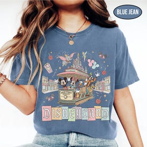 May include: Blue jean colored t-shirt featuring a Disneyland design. The graphic includes Mickey Mouse, Minnie Mouse, Donald Duck, and Goofy riding a Main Street Trolley. The Disneyland logo is displayed in a colorful, block letter style.