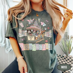 May include: Moss green t-shirt featuring a Main Street Trolley with Mickey, Minnie, Donald, and Goofy. The shirt has a Disneyland graphic with Dumbo, fireworks, and popcorn. The text "DISNEYLAND" is in colorful block letters.