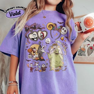 May include: A violet Comfort Colors t-shirt featuring a colorful graphic of characters from The Nightmare Before Christmas. The design includes Jack Skellington, Sally, Oogie Boogie, and other elements from the movie. The shirt has a relaxed fit.