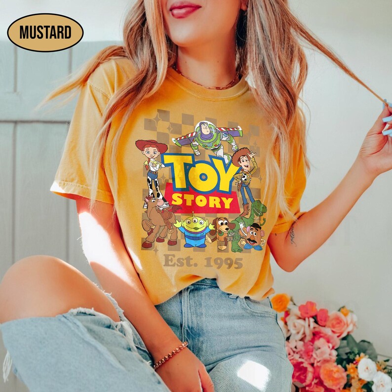 May include: Mustard-colored t-shirt featuring a graphic of Toy Story characters, including Woody, Buzz Lightyear, and others. The words "TOY STORY" are in a bold, colorful font, with "Est. 1995" below. The shirt has a classic crew neck.