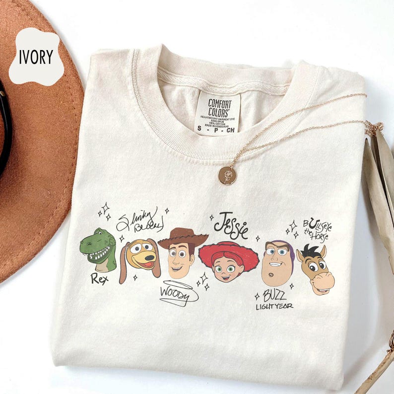 May include: An ivory short-sleeved t-shirt featuring a graphic print of six cartoon character heads from Toy Story, including Rex, Slinky Dog, Woody, Jessie, Buzz Lightyear, and Bullseye, with their names written in black script text.