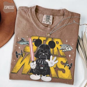 Puede incluir: Camiseta color espresso con un dibujo animado de Darth Vader con orejas de Mickey sosteniendo un sable de luz. La camiseta tiene las palabras "STAR WARS" en amarillo, junto con ilustraciones de naves espaciales y la etiqueta de la marca Comfort Colors.