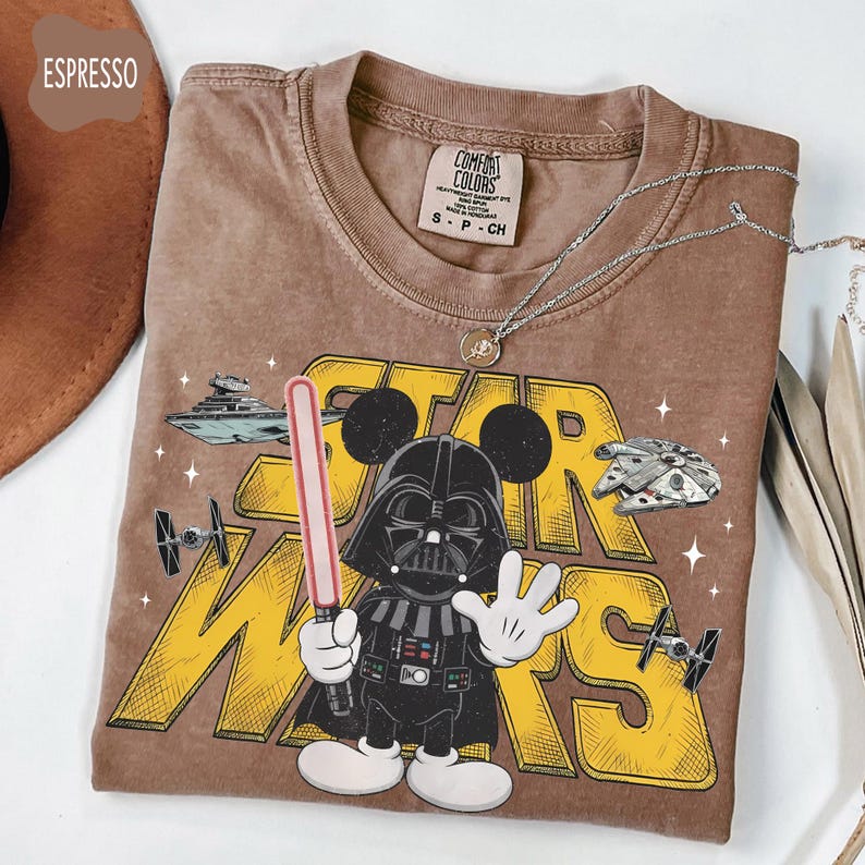 Puede incluir: Camiseta color espresso con un dibujo animado de Darth Vader con orejas de Mickey sosteniendo un sable de luz. La camiseta tiene las palabras "STAR WARS" en amarillo, junto con ilustraciones de naves espaciales y la etiqueta de la marca Comfort Colors.