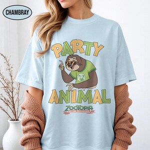 May include: Light blue t-shirt with the words "PARTY ANIMAL" in green and orange block letters. A cartoon sloth wearing sunglasses and a green shirt is in the center. The text "ZOOTOPIA BETTER ZOOGETHER" is below the sloth.