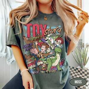 Puede incluir: Camiseta verde musgo con un gr&aacute;fico colorido de Toy Story, con Woody, Buzz Lightyear y Rex. El texto "TOY STORY" est&aacute; sobre los personajes, con "SINCE 1995" debajo. La camiseta tiene mangas cortas.