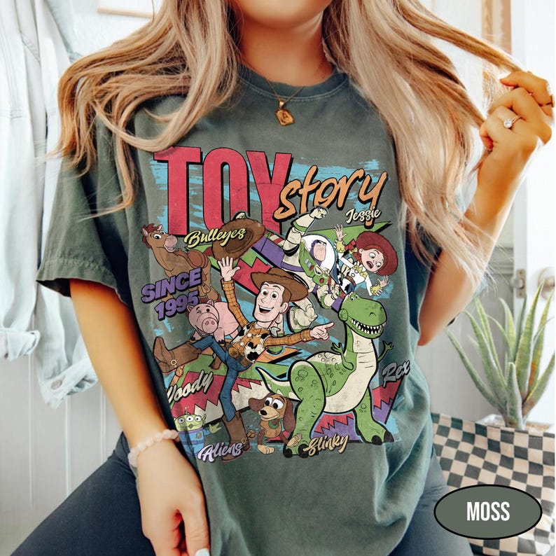 Puede incluir: Camiseta verde musgo con un gr&aacute;fico colorido de Toy Story, con Woody, Buzz Lightyear y Rex. El texto "TOY STORY" est&aacute; sobre los personajes, con "SINCE 1995" debajo. La camiseta tiene mangas cortas.