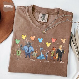 Puede incluir: Camiseta color espresso con personajes de dibujos animados sosteniendo globos con forma de Mickey Mouse. La camiseta tiene la etiqueta de la marca Comfort Colors y un collar plateado. Personajes: Flash, Bogo, Gary, Judy Hopps y Mayor.