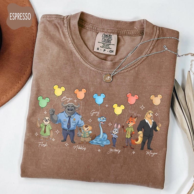 Puede incluir: Camiseta color espresso con personajes de dibujos animados sosteniendo globos con forma de Mickey Mouse. La camiseta tiene la etiqueta de la marca Comfort Colors y un collar plateado. Personajes: Flash, Bogo, Gary, Judy Hopps y Mayor.