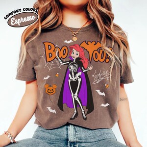 May include: An espresso-colored t-shirt featuring a graphic of a red-haired character in a skeleton costume with a purple-lined black cape. The text reads Boo You! with a spiderweb, bats, candy corn, and a pumpkin icon. The Comfort Colors logo is visible.